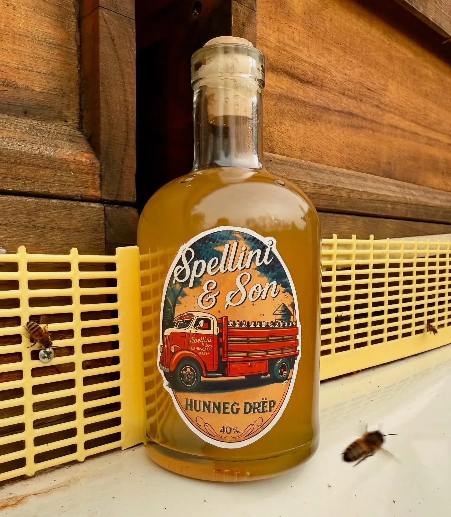 Bouteille de goutte de miel étiquetée Spellini & Son, posée devant un treillis de bois, une abeille à côté